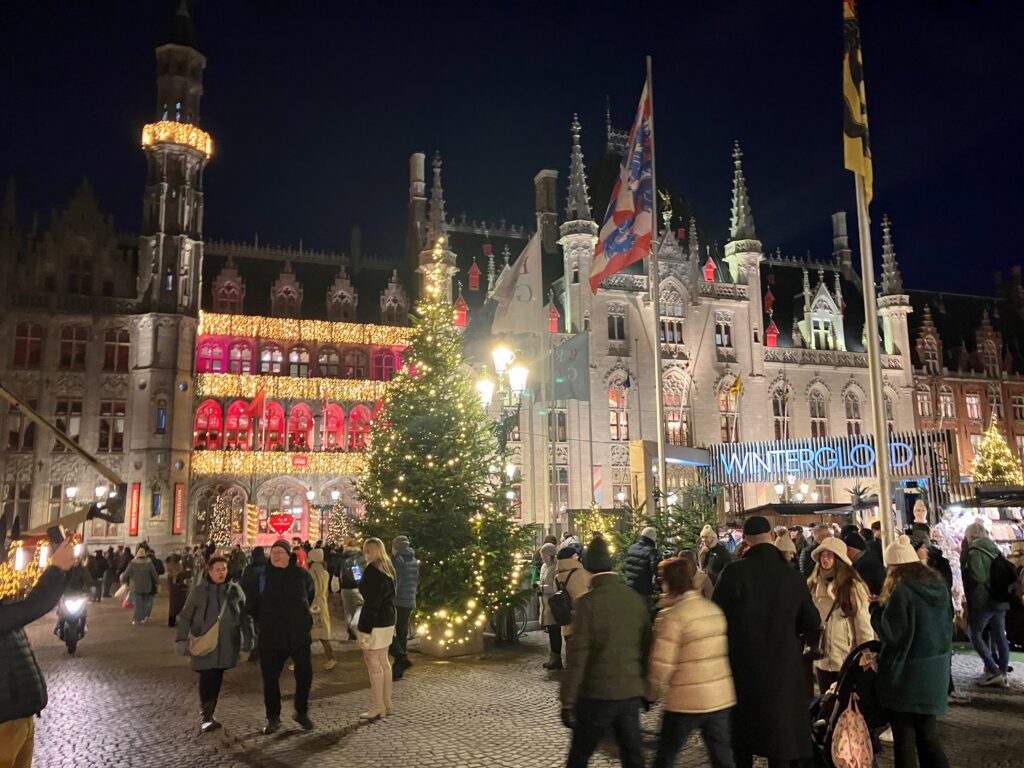 Bruges winter market glowing at night with festive lights, historic Flemish architecture, Christmas trees, and joyful crowds—captured for Amore Travel Designs, a Fort Collins luxury travel advisor crafting bespoke European holiday journeys.