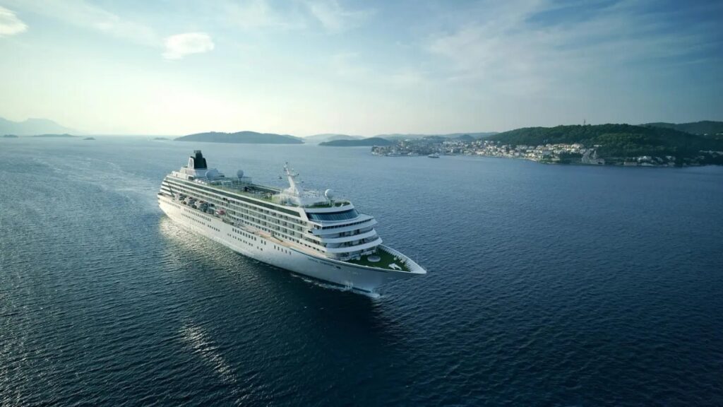 Crystal Serenity luxury cruise ship sailing through calm blue waters near a European coastline, showcasing the elegant lifestyle and refined cruising experience curated by Amore Travel Designs, Fort Collins’ luxury travel advisor.