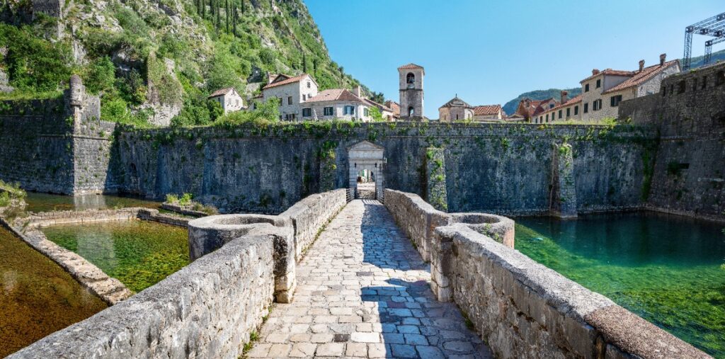 A sunlit cobblestone bridge leads through the medieval gate of Kotor, Montenegro, flanked by crystal-clear moat waters — one of Europe's most magical hidden destinations, as featured by Cathy at Amore Travel Designs.