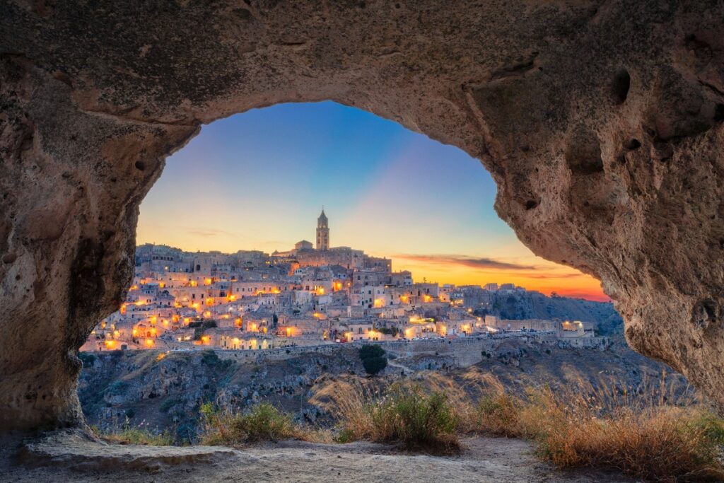 Matera, Italy lit up at dusk, viewed through the opening of an ancient cave — one of Europe's most extraordinary hidden destinations, as featured by Fort Collins luxury travel advisor Cathy at Amore Travel Designs