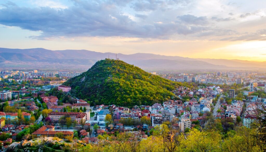 Panoramic sunset view of Plovdiv, Bulgaria, with the city's iconic tree-covered hill rising from a sea of terracotta rooftops — one of Europe's oldest and most overlooked luxury travel destinations, featured by Amore Travel Designs.