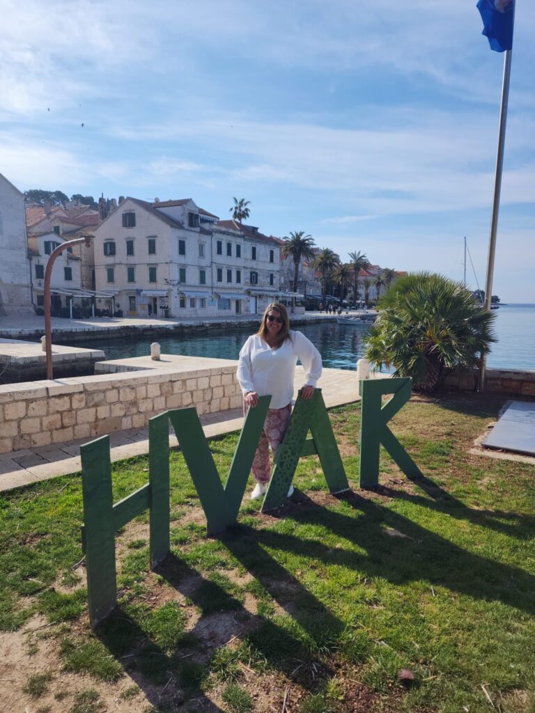 Fort Collins-based luxury travel advisor posing by the HVAR sign in a waterfront park, Hvar, Croatia, with harbor scenery behind. 🧭