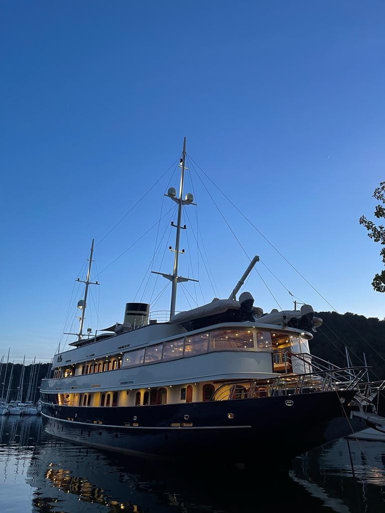 Fort Collins-based luxury travel advisor’s yacht for the week moored in a marina at blue hour, featuring a navy hull, cream decks, and warm interior glow.