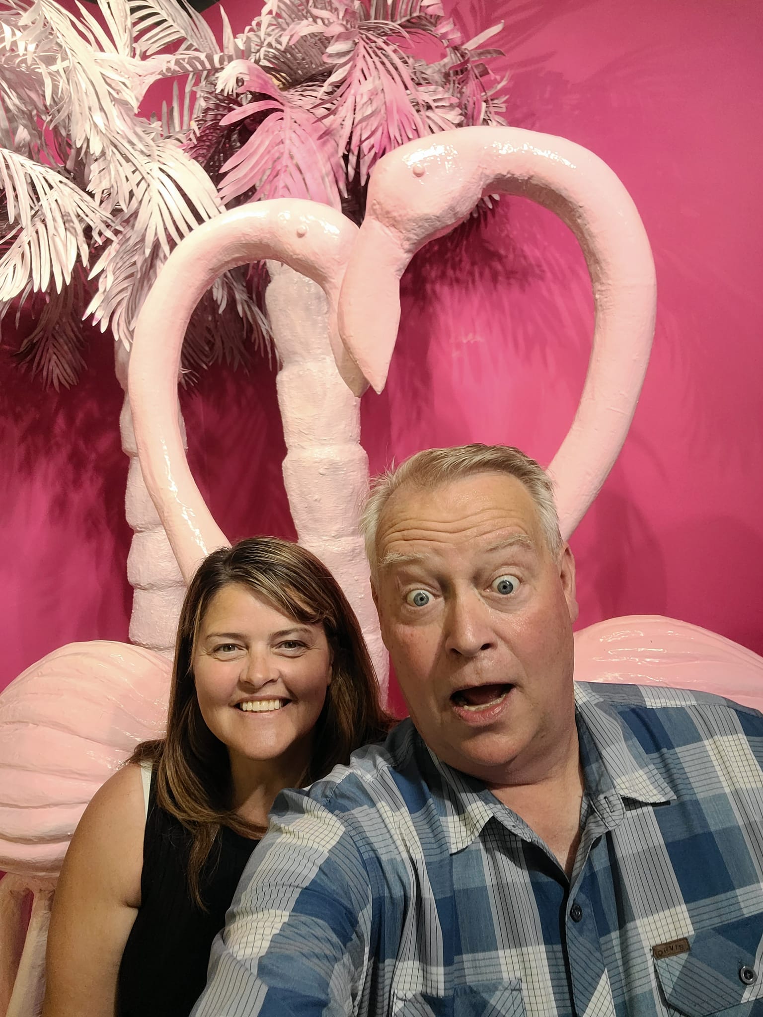 Fort Collins-based luxury travel advisor and husband in a tropical flamingo backdrop, with a magenta wall and palm fronds behind them. In the Dubrovnik selfie museum
