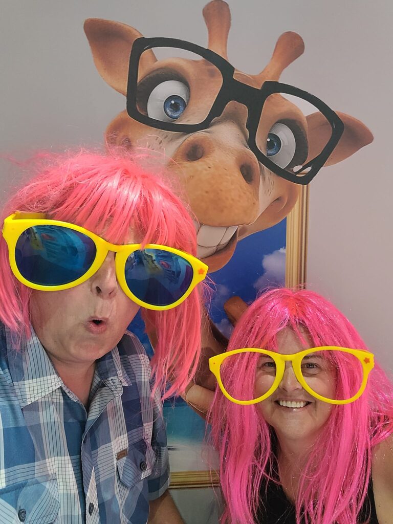 Fort Collins-based luxury travel advisor and husband in Dubrovnik selfie museum, pink wigs, yellow frames, with a giraffe prop and blue-toned wall behind them.